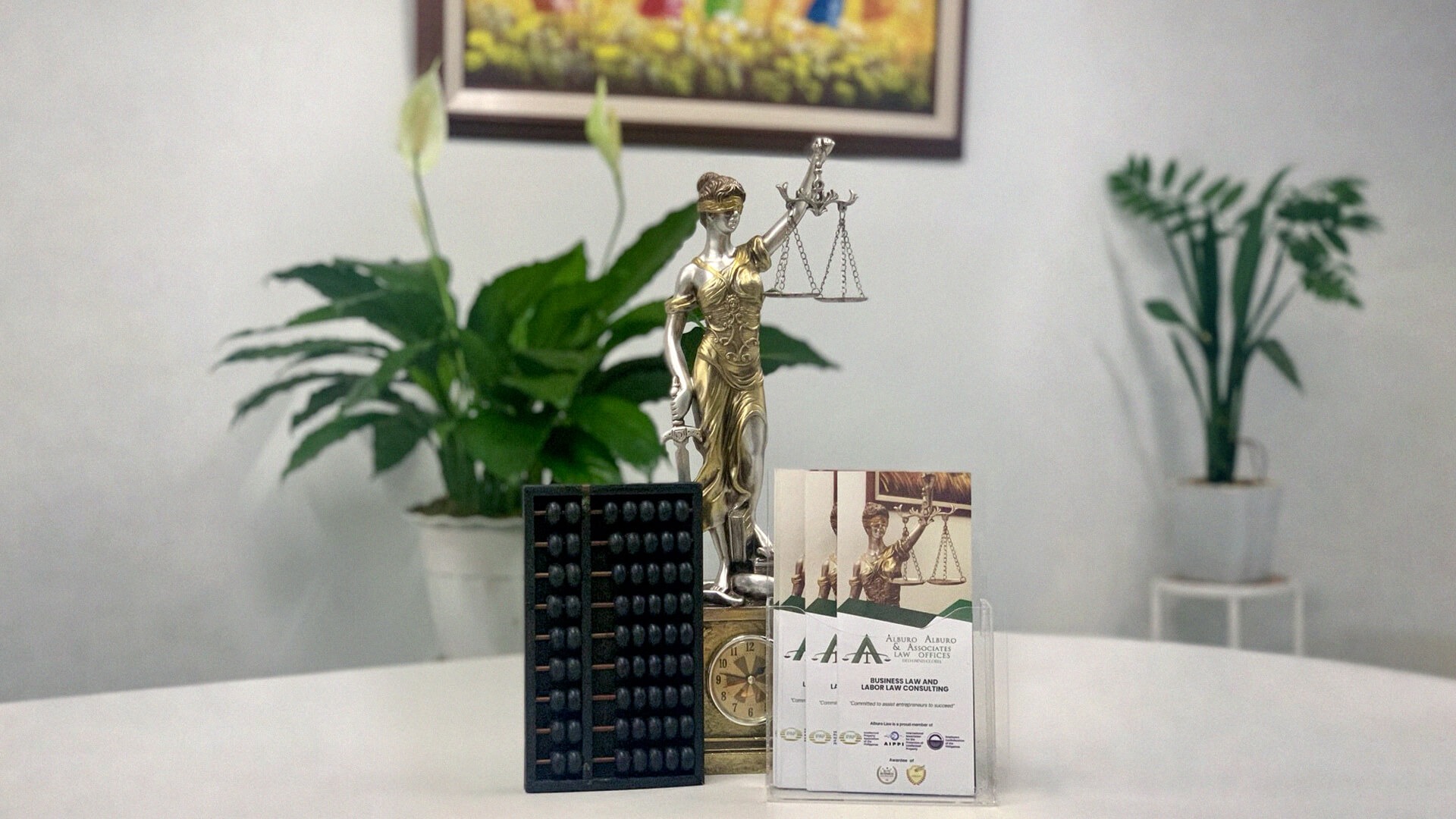 A professional legal setting featuring a Lady Justice statue, a traditional abacus, and Alburo Law brochures on a clean white desk, with lush green plants in the background.