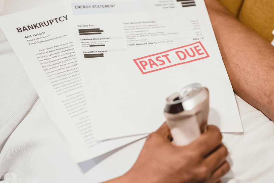 Person holding a crushed soda can while looking at an energy bill with a "PAST DUE" stamp and a "BANKRUPTCY" notice, illustrating financial debt and collection issues.