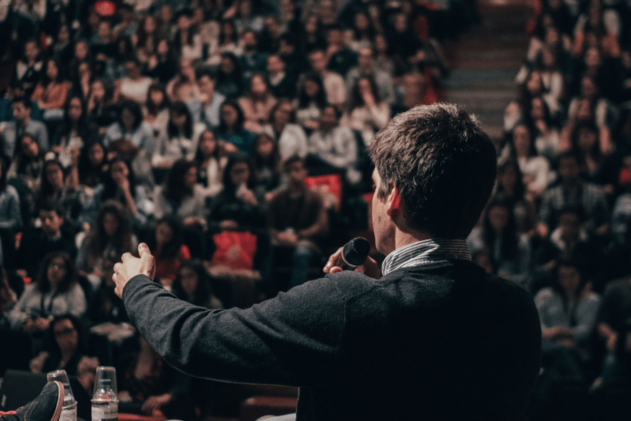 Back view of a public official speaking into a microphone before a large crowded audience in an auditorium, representing civil service and administrative law.