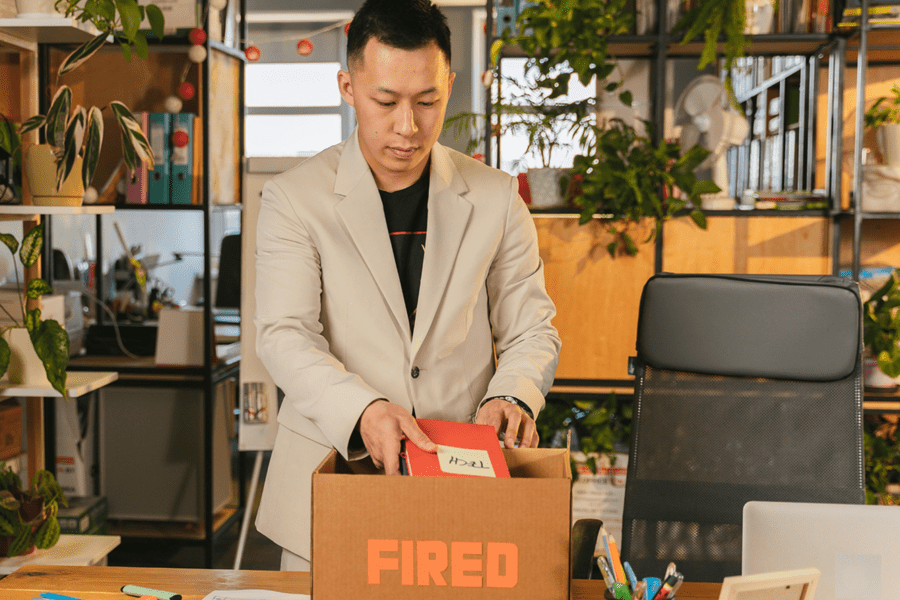 A man in a beige blazer packing a box with a "FIRED" label in a modern office, representing termination of employment and labor law disputes.