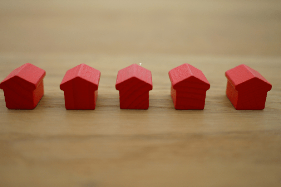 A row of five small red wooden toy houses on a light wood surface, representing real estate, property ownership, and land titles.