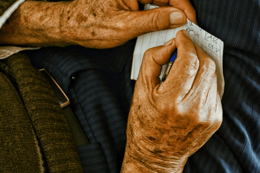 Close-up of an elderly person's weathered hands writing on a small piece of paper with a blue pen, symbolizing the drafting of a last will and testament.
