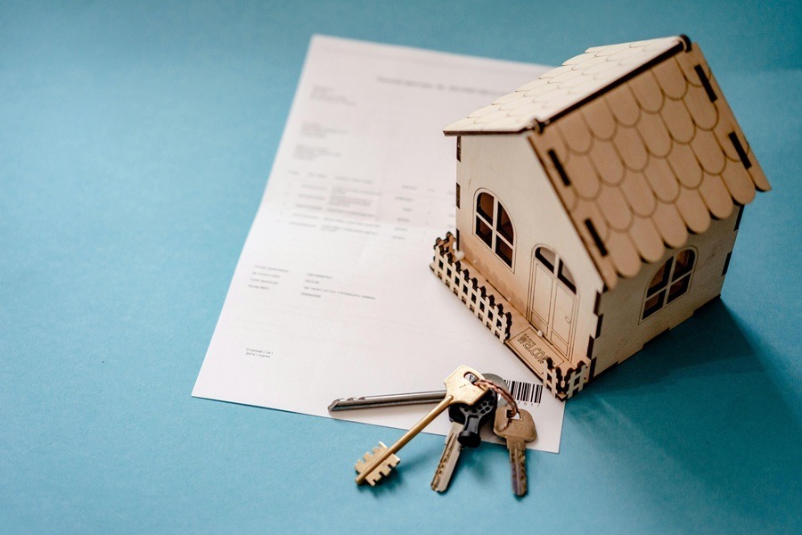 A small wooden model house sits on top of a legal document, accompanied by a set of keys resting on a blue background.