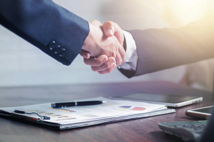 A professional handshake between two individuals in a formal office setting, symbolizing a business agreement.