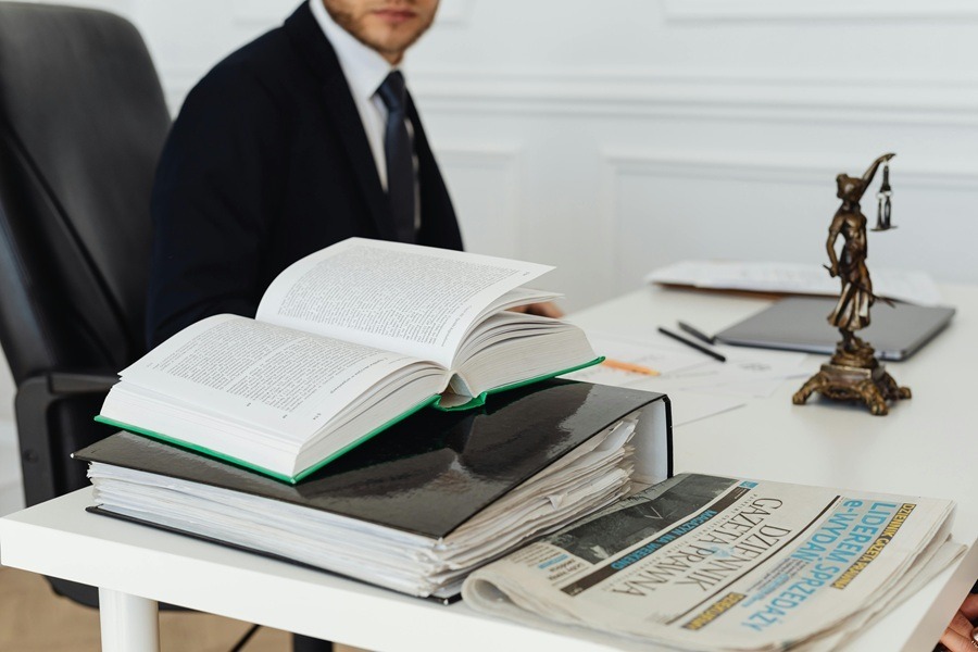 A professional office scene featuring a person in a suit, a stack of legal documents with an open book, and a bronze statuette of Lady Justice on a white desk.